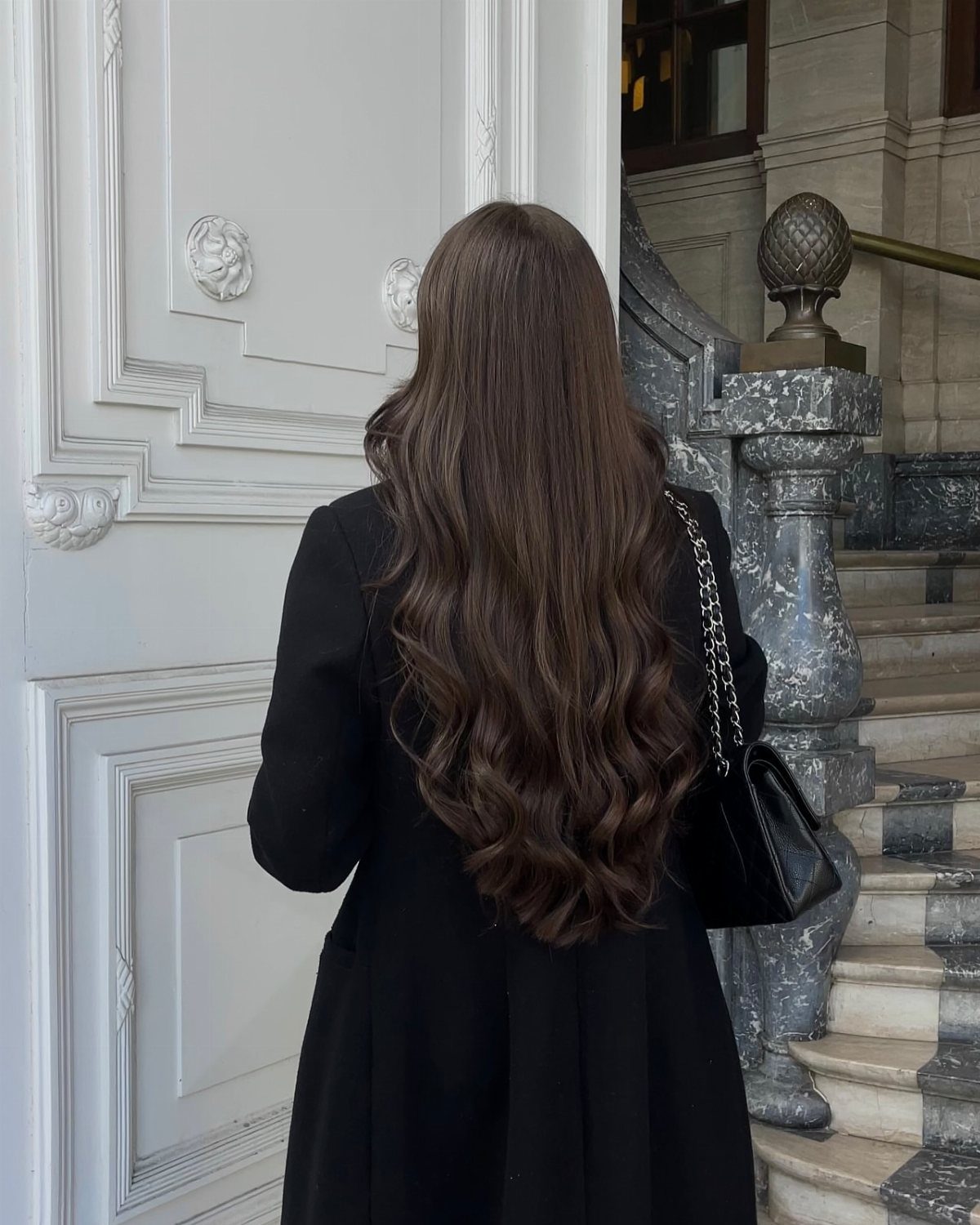 Long glossy dark brunette waves fall down the back of a person in a black coat
