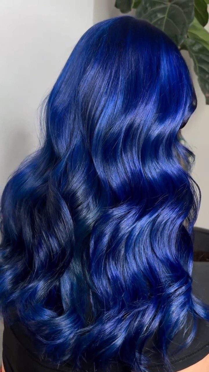 Long royal-blue hair falls in glossy curls against a pale wall