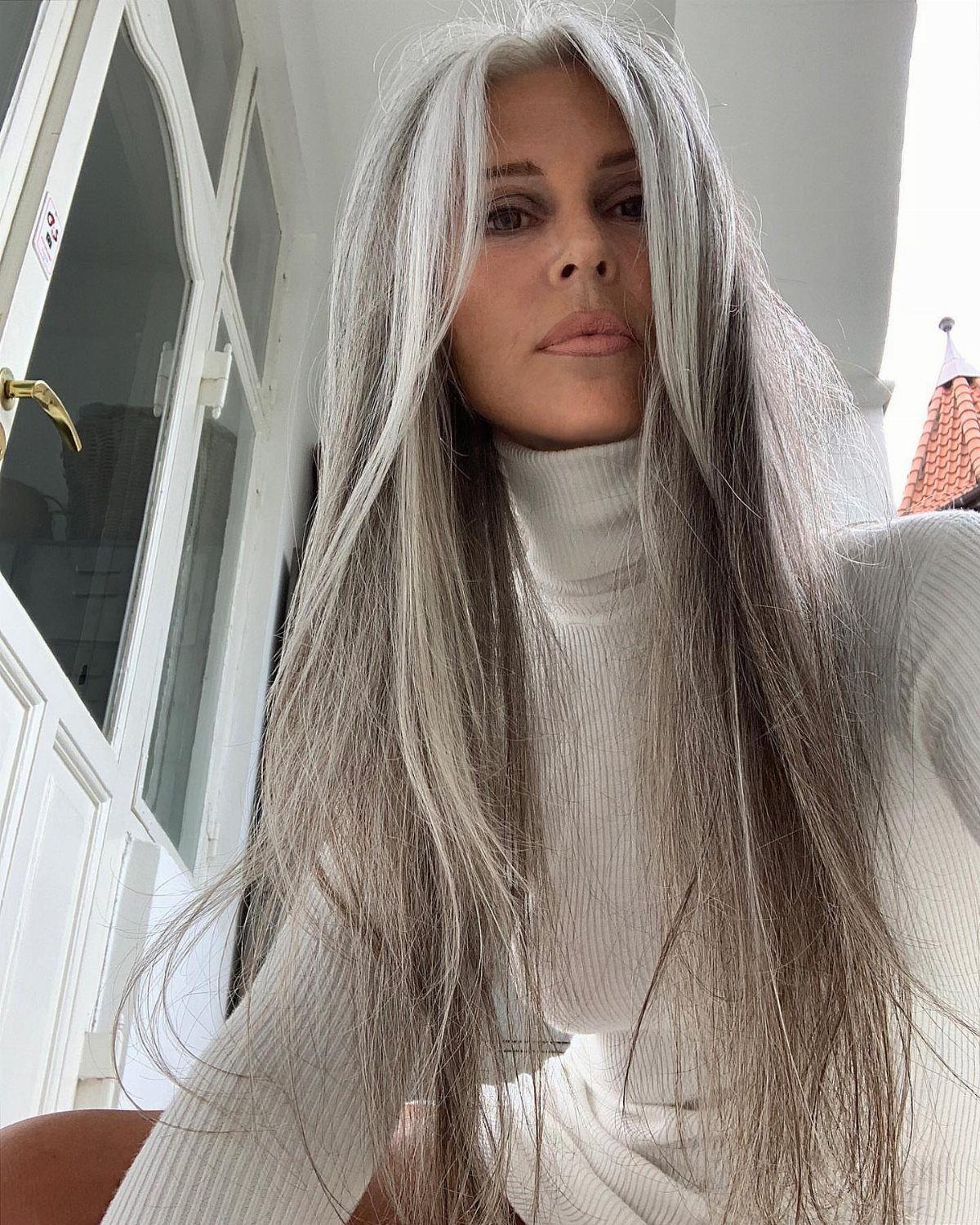 Woman with long silver-gray hair and darker roots wearing a white turtleneck
