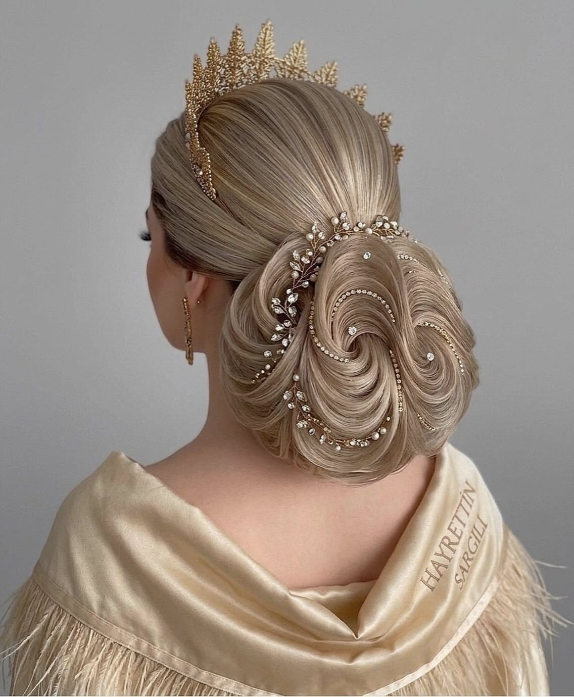 Blonde low chignon styled in curled loops with crystal pins and a gold crown