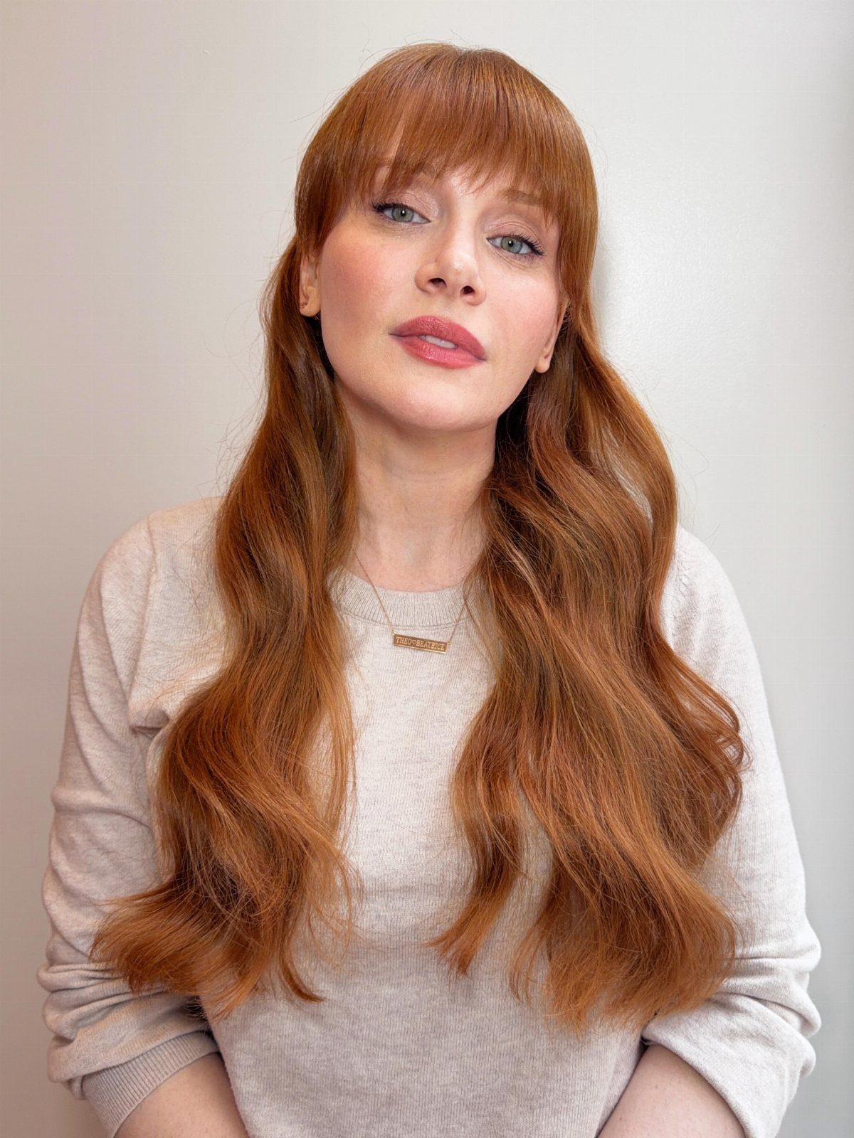 Woman with long wavy strawberry-brown hair and bangs against a light wall