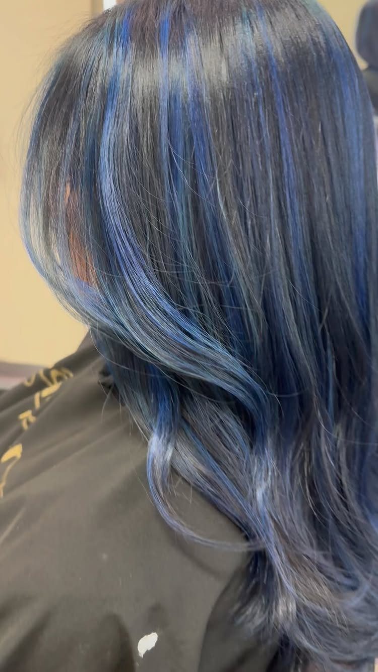 Wavy dark hair with blue highlights drapes over a salon cape