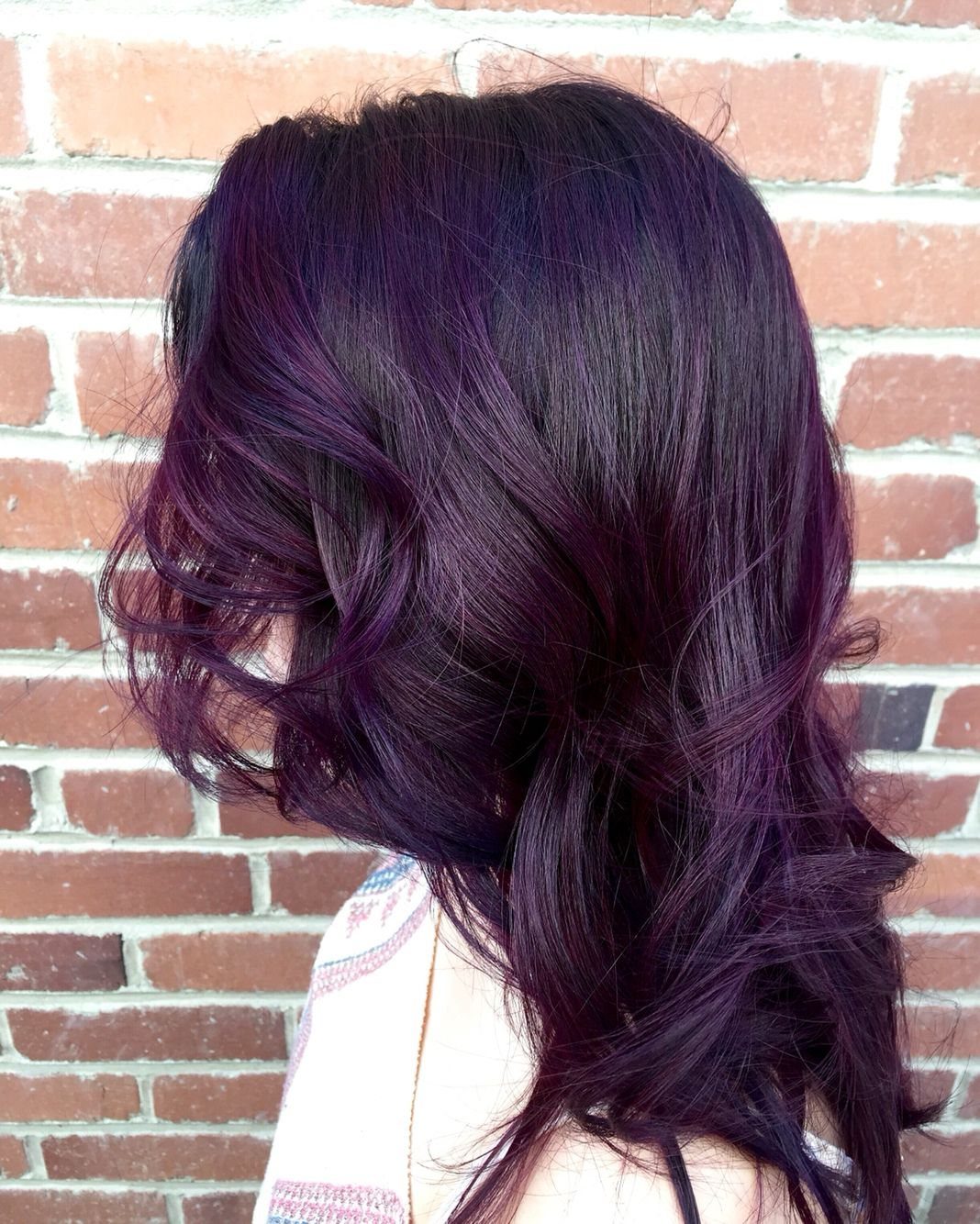Glossy plum-purple layered hair falls in soft waves against a brick wall.