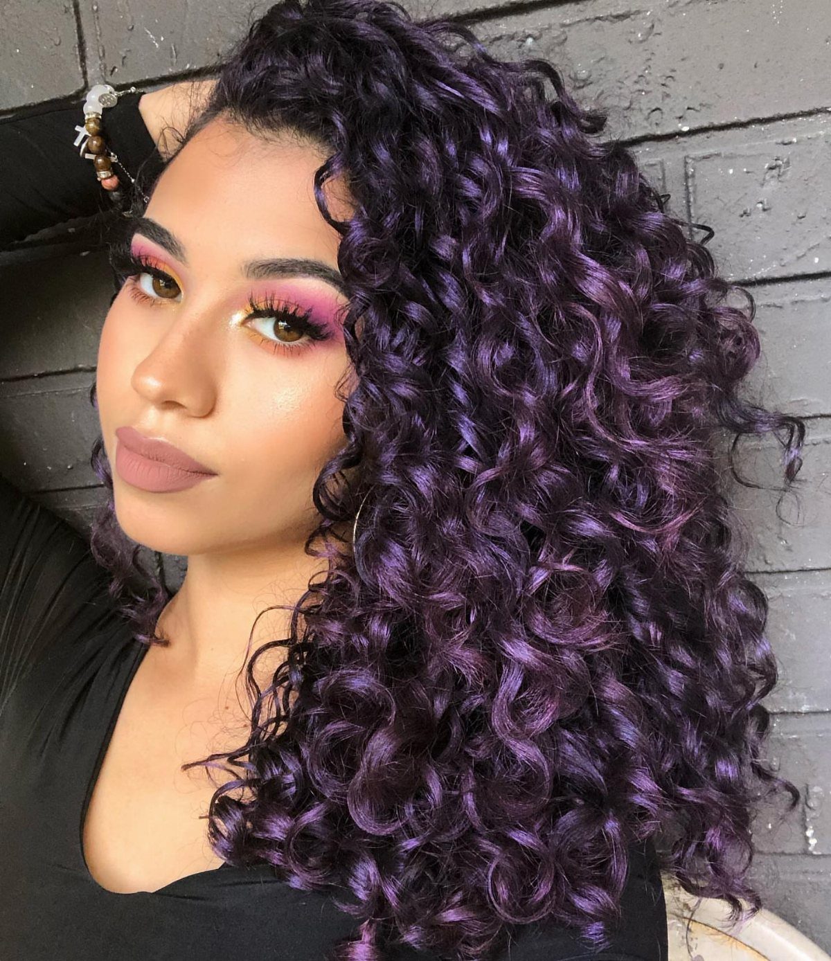 Woman with voluminous dark purple curls posing against a gray wooden wall