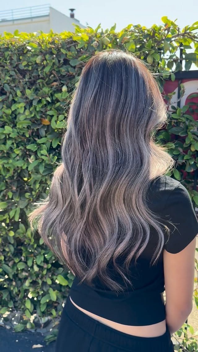 Long wavy brunette hair shows soft, freehand balayage highlights against a leafy hedge