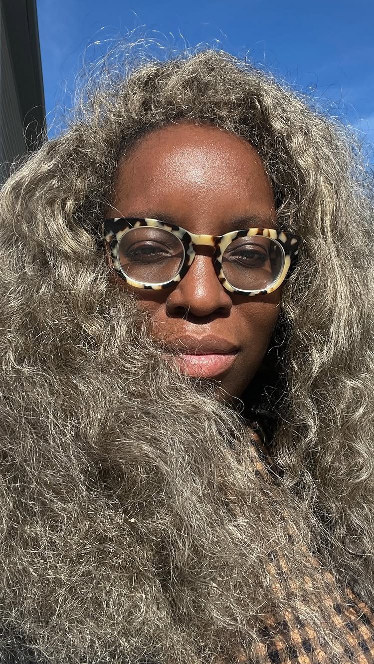 Curly dark hair with silvery-gray highlights frames a person wearing tortoiseshell glasses.