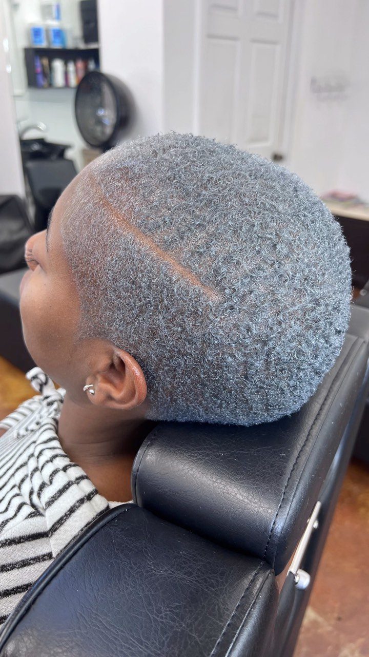 Short gray curls with darker roots and a shaved line in a salon chair