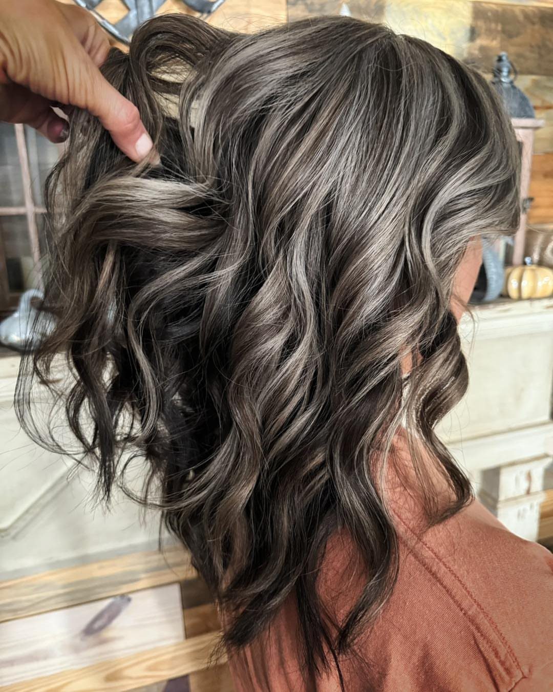 Wavy ash-brown hair with silver-gray strands lifted by a hand in a salon
