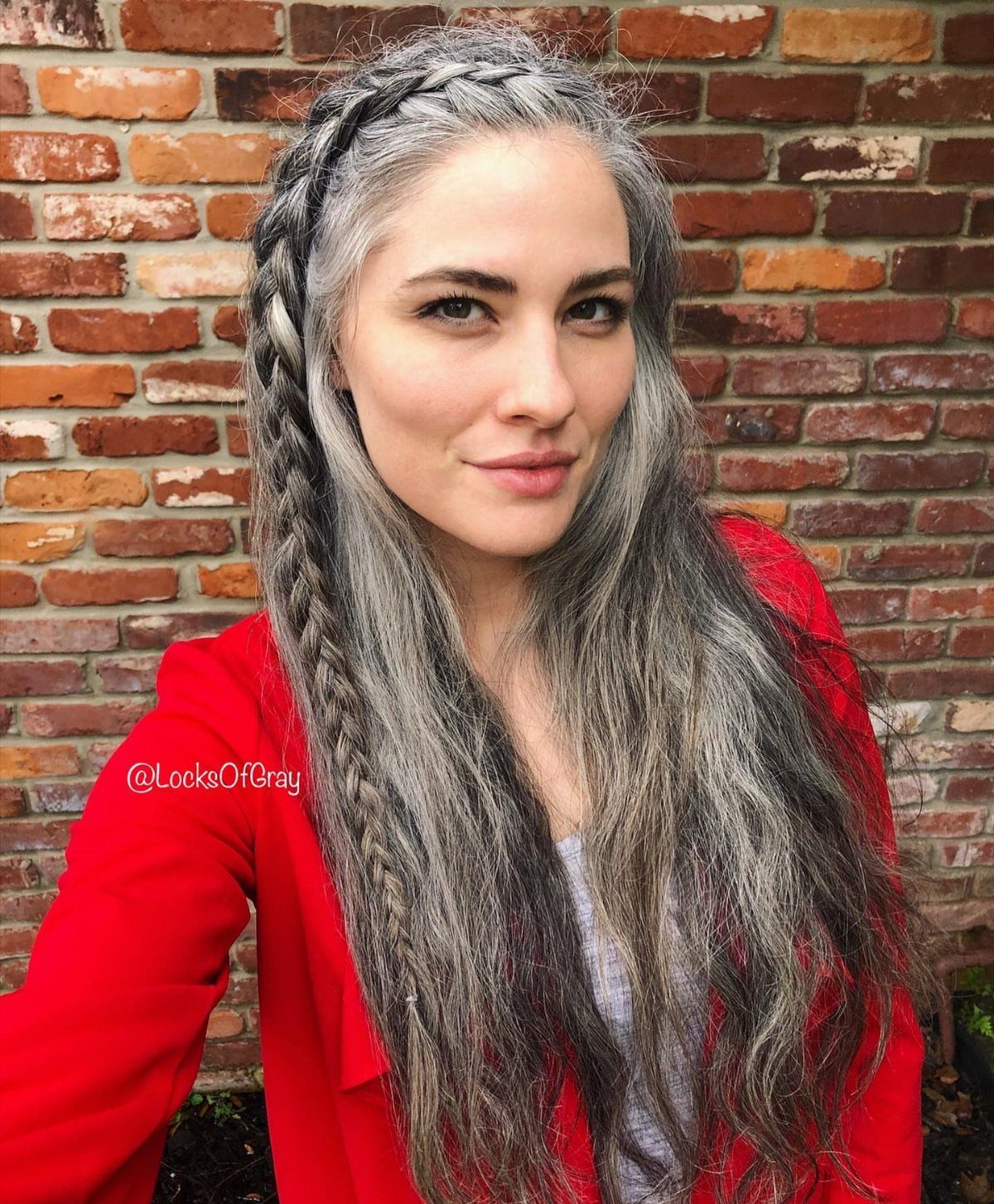 Woman with long grey hair and a crown braid in front of a brick wall