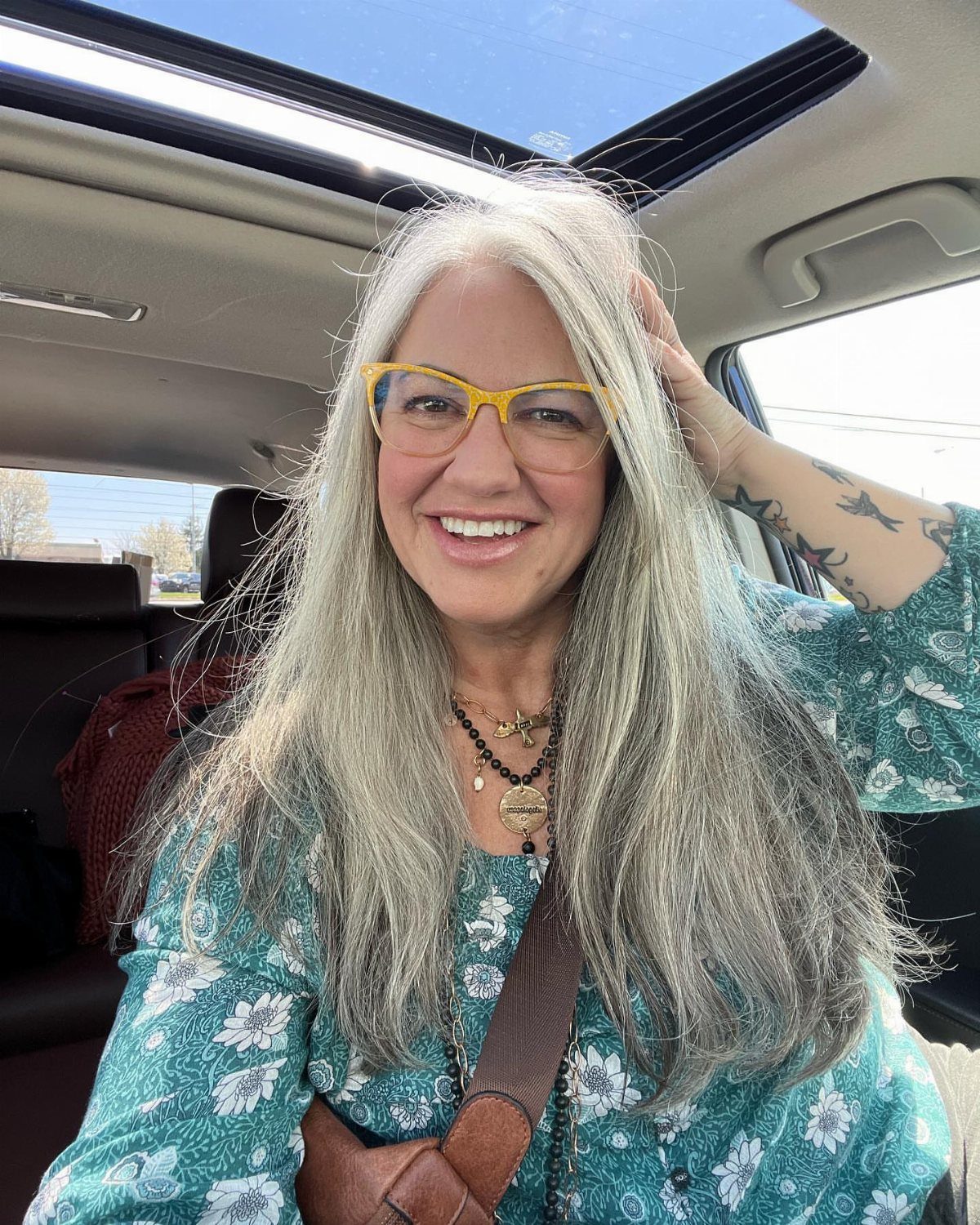 Woman with long grey hair and yellow glasses smiles in a car