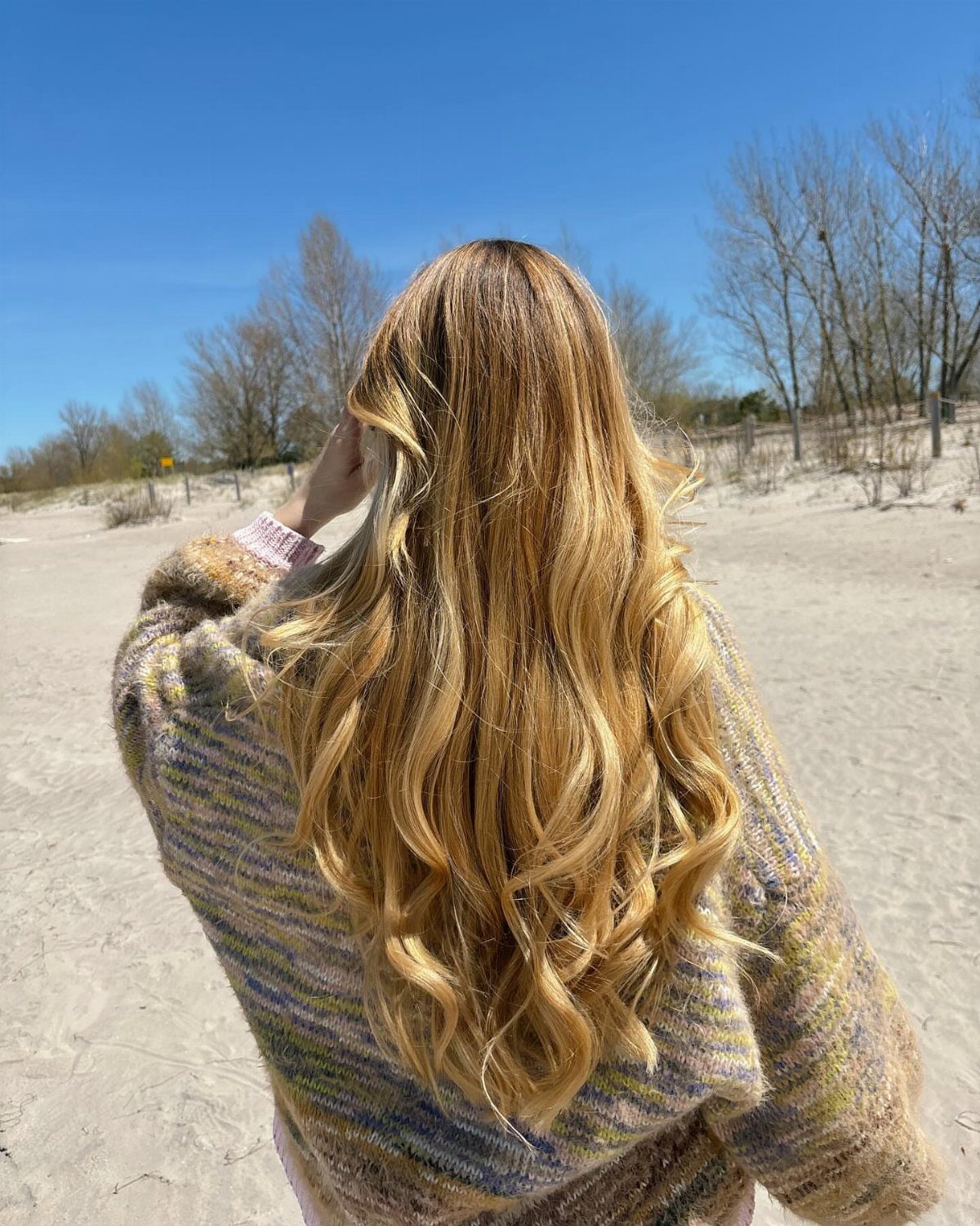Long golden-blonde waves fall over a knit sweater on a sunny beach