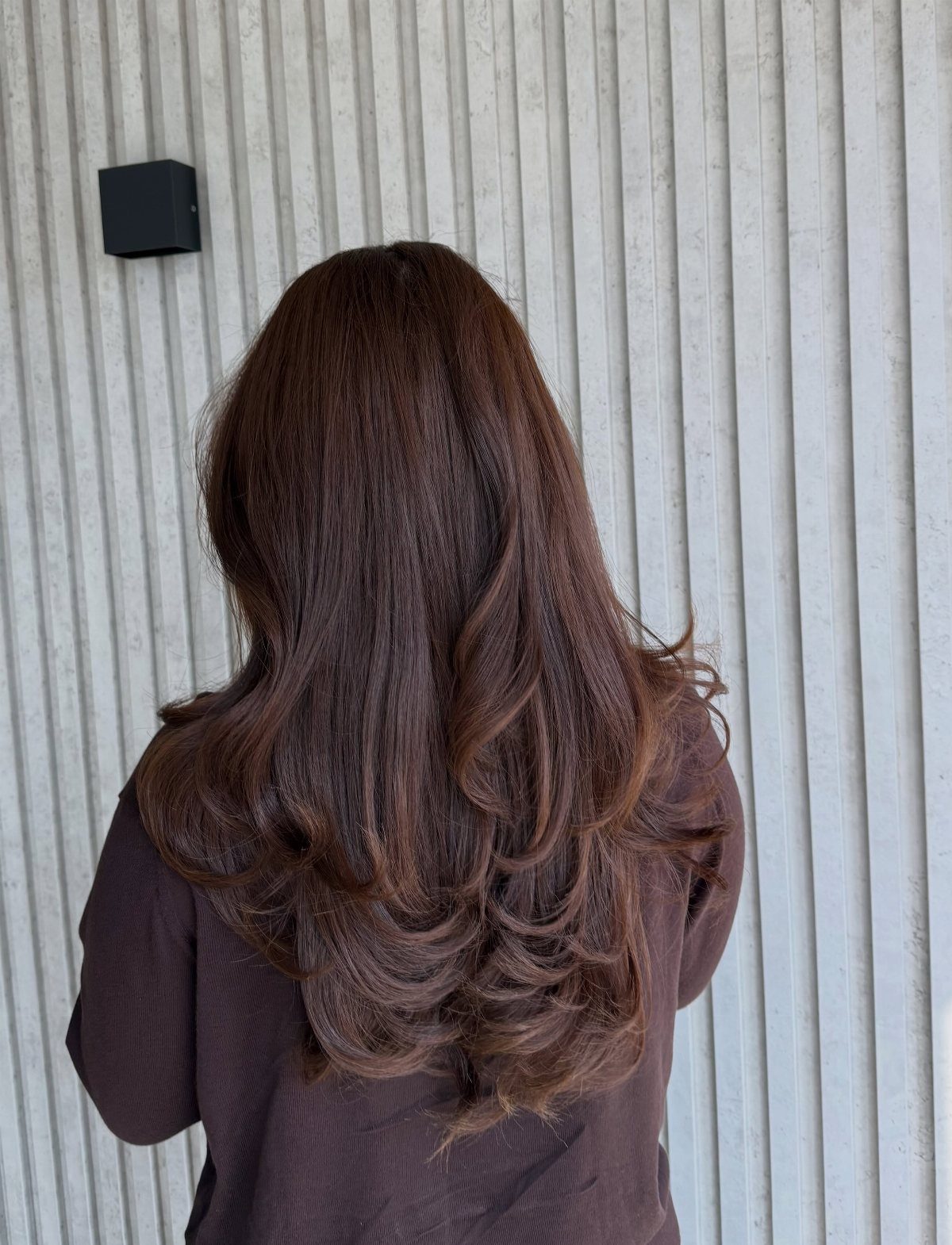 Long mocha-brown curls fall over a dark mauve top against a ribbed wall
