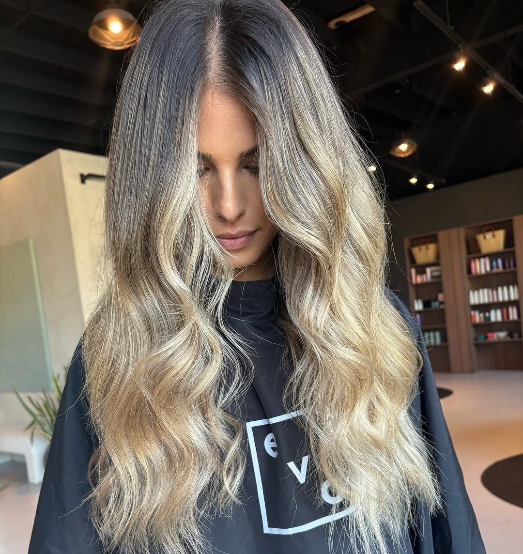 Long wavy beige blonde hair with darker roots, styled on a woman in a salon