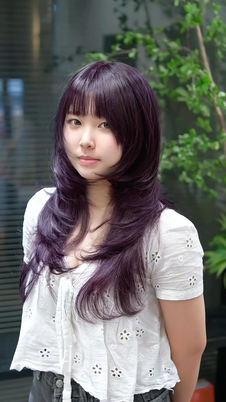 Woman showing long, layered violet hair with bangs against a glass building