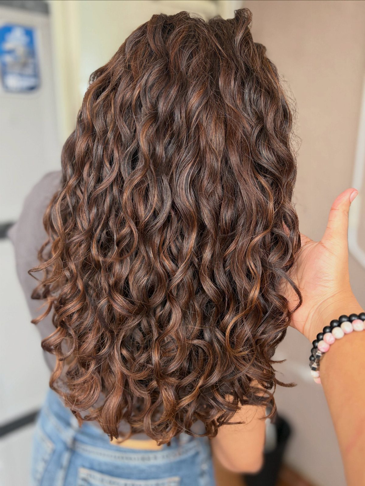 Curly brown hair with lighter highlights shown from behind in a salon