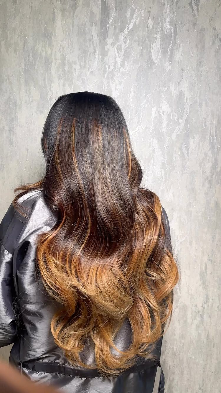 Long brunette hair blends into caramel and honey-colored ends against a textured gray wall.