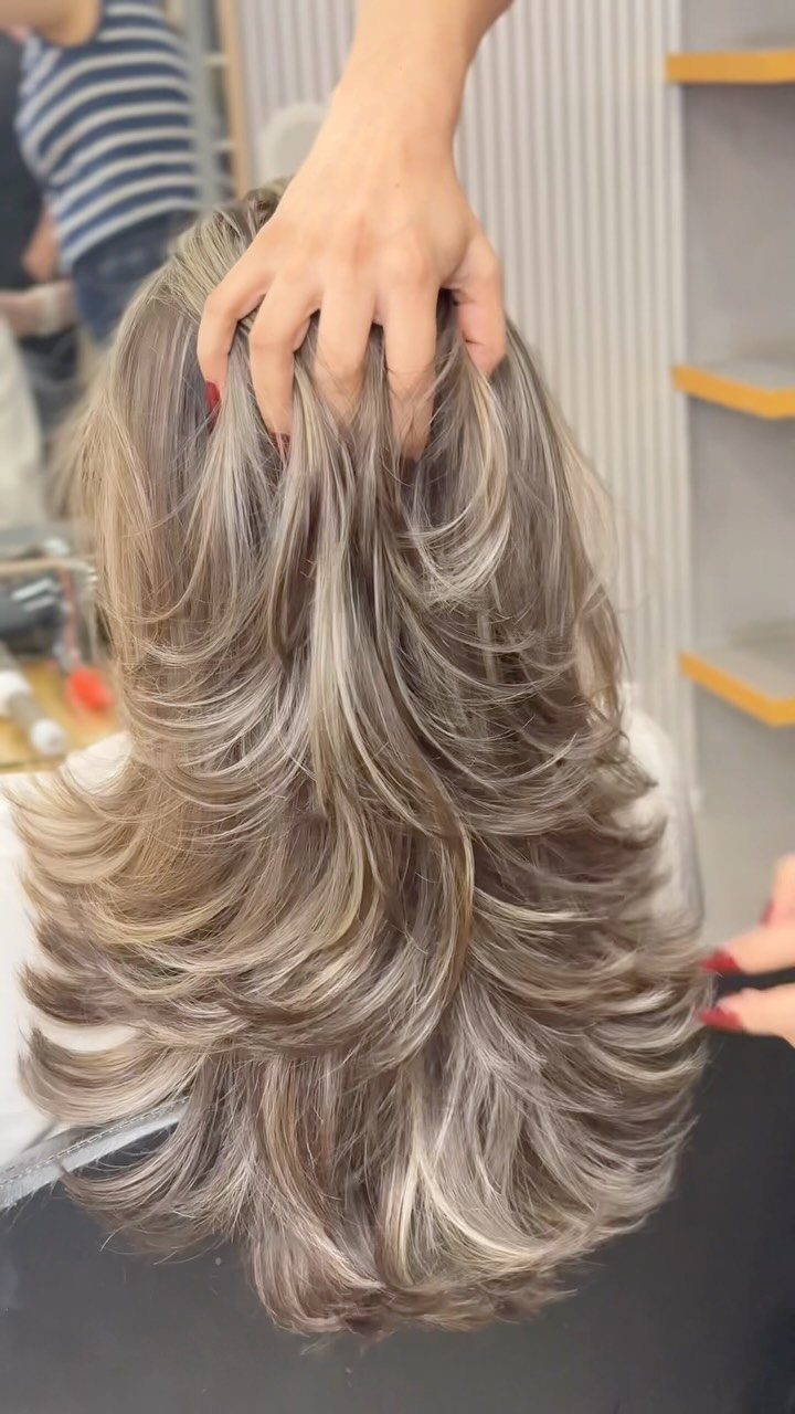 Layered gray-brown hair with pale blonde highlights parted by a hand in a salon