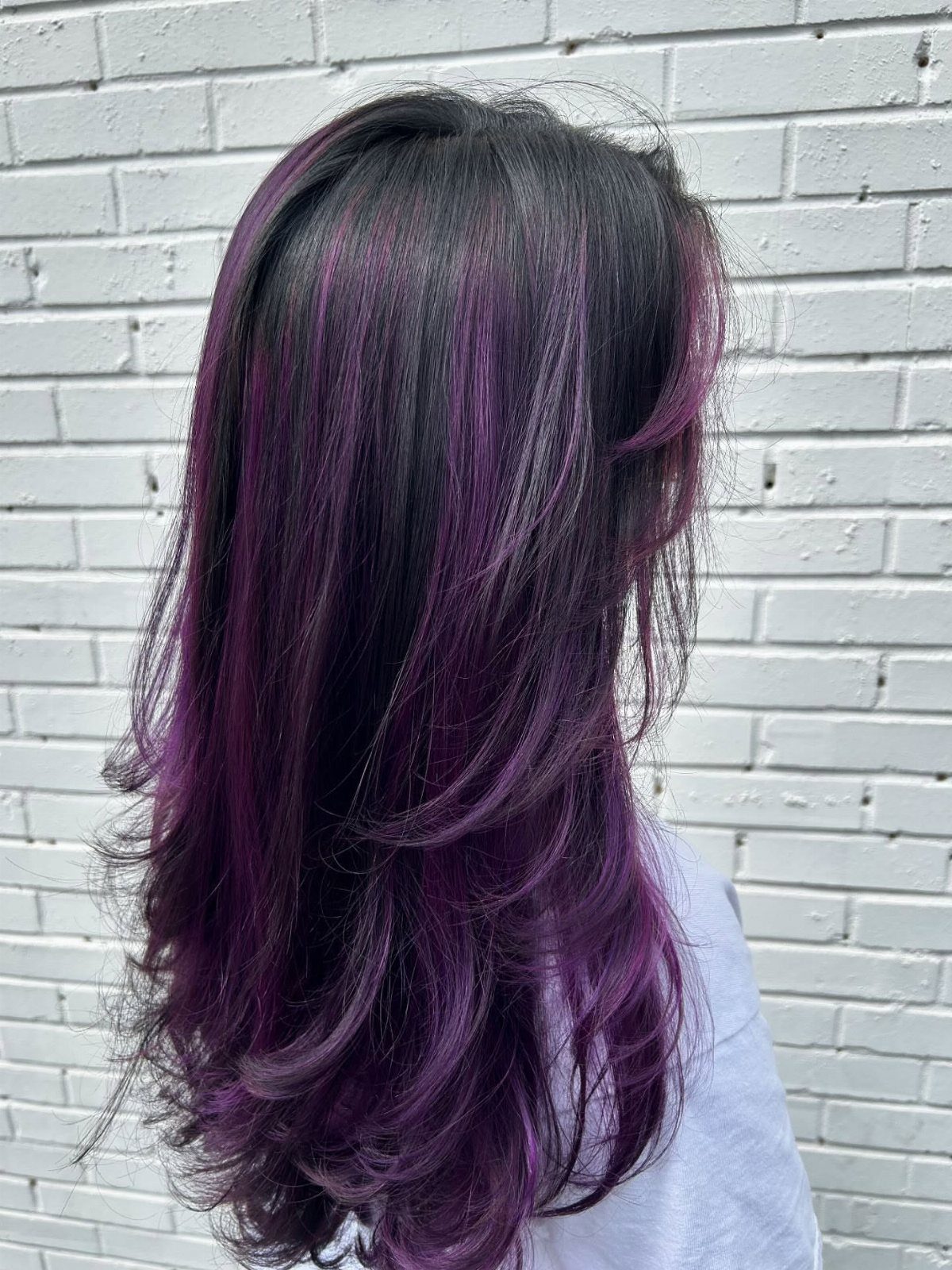 Long dark hair with purple highlights and curled layers against white brick