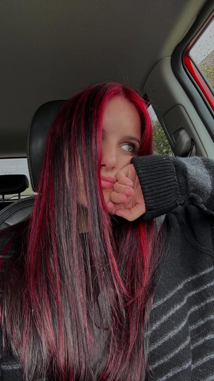 Long dark hair with vivid red highlights frames a person seated inside a car.