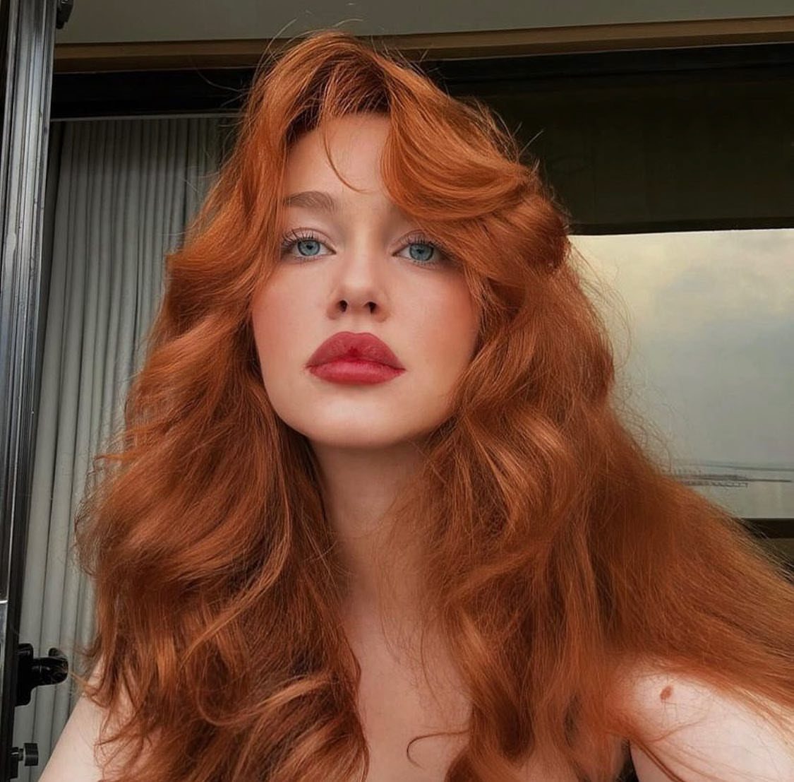 Woman with long, voluminous copper-red hair and blue eyes posing outdoors