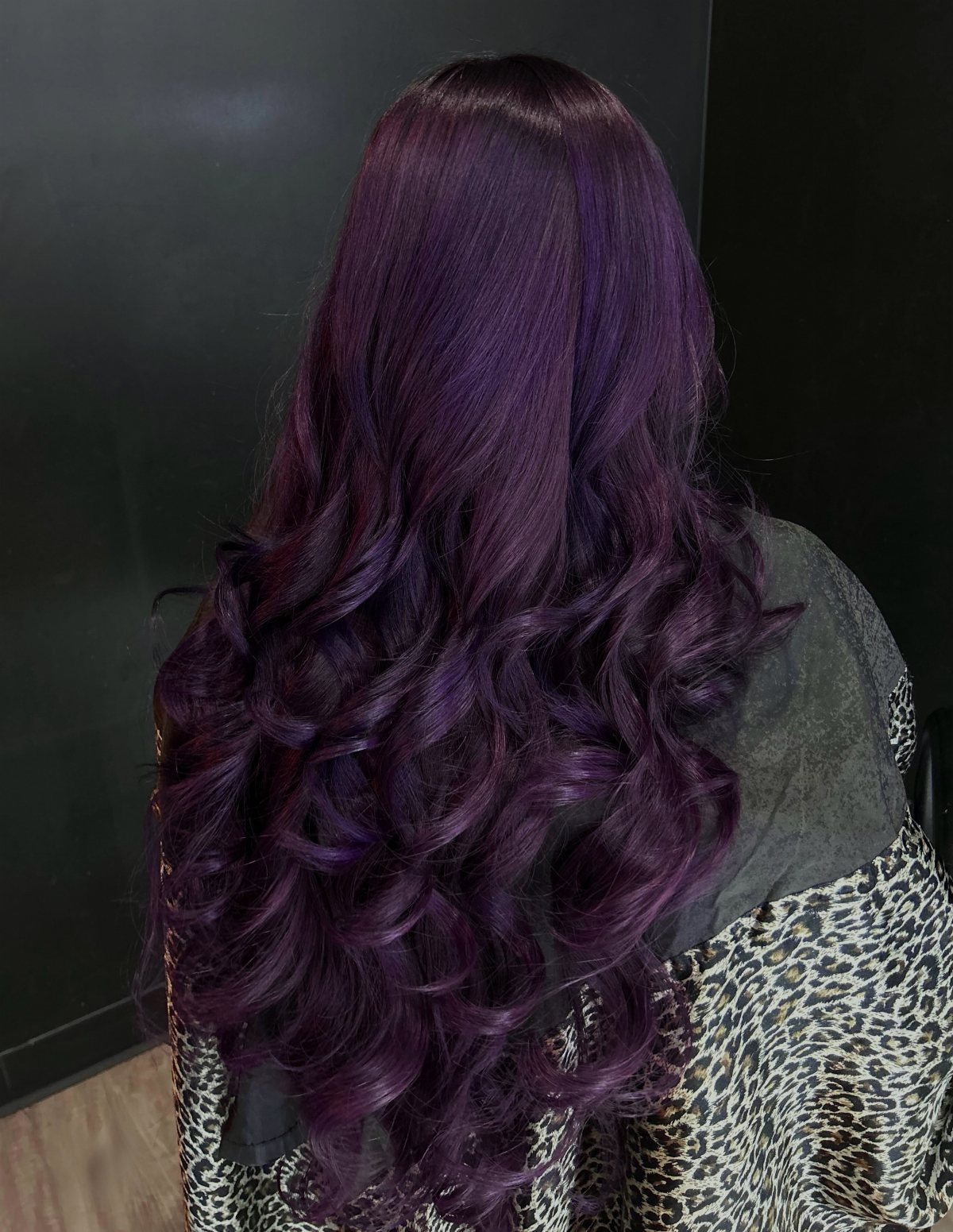 Long curled violet hair with dark roots viewed from behind against a dark wall
