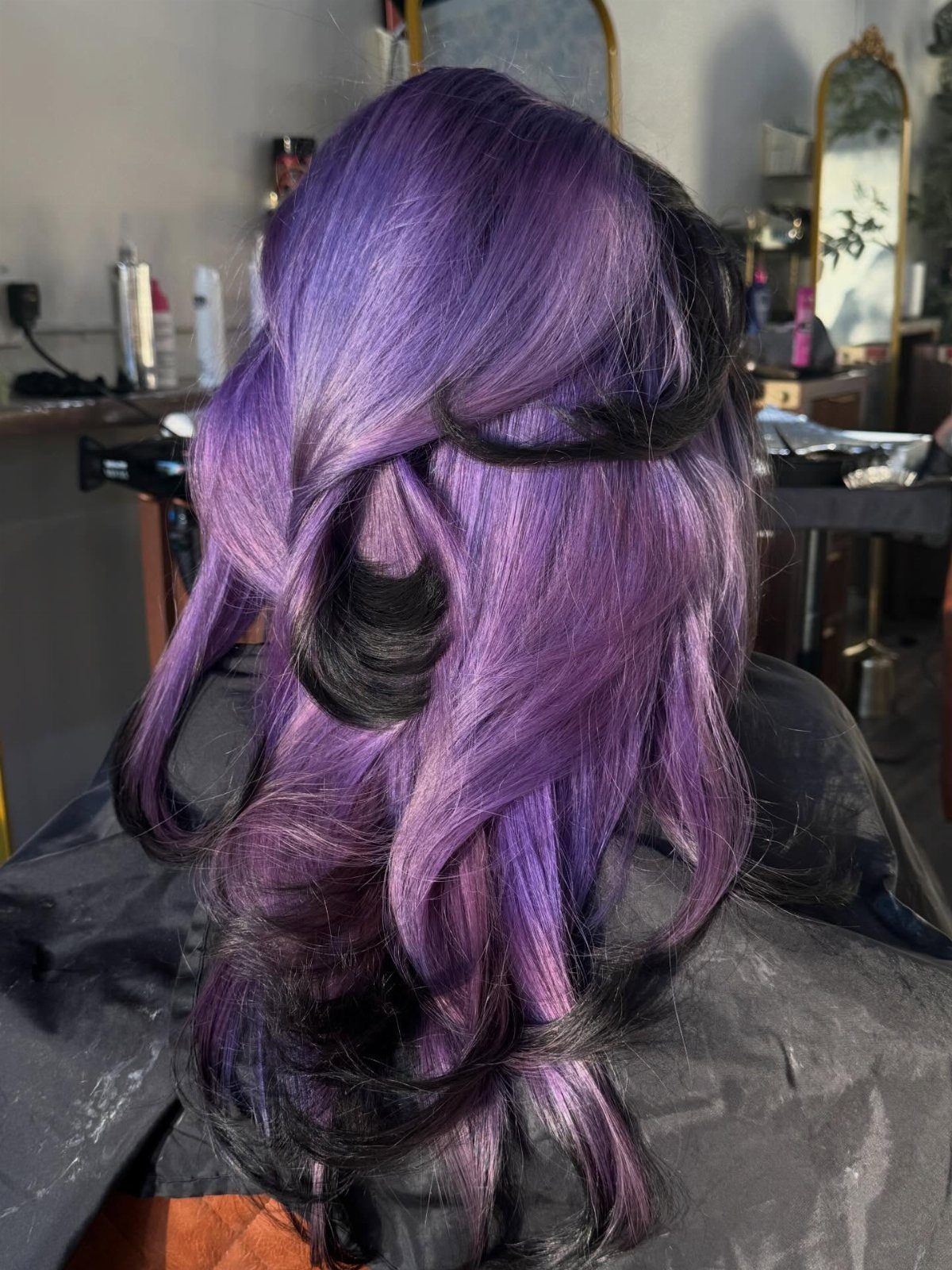 Long lavender-purple curls with dark sections styled over a salon cape