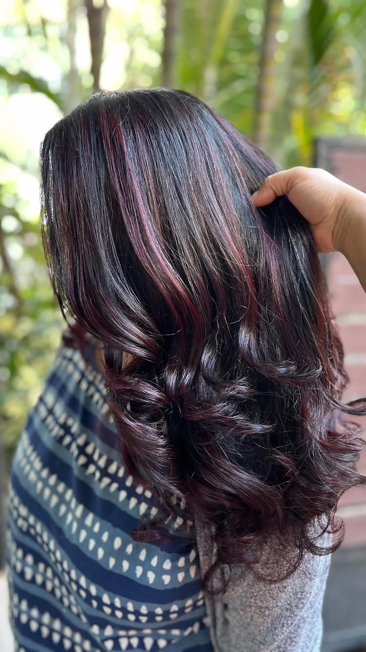 Dark curled hair with burgundy streaks is lifted by a hand outdoors