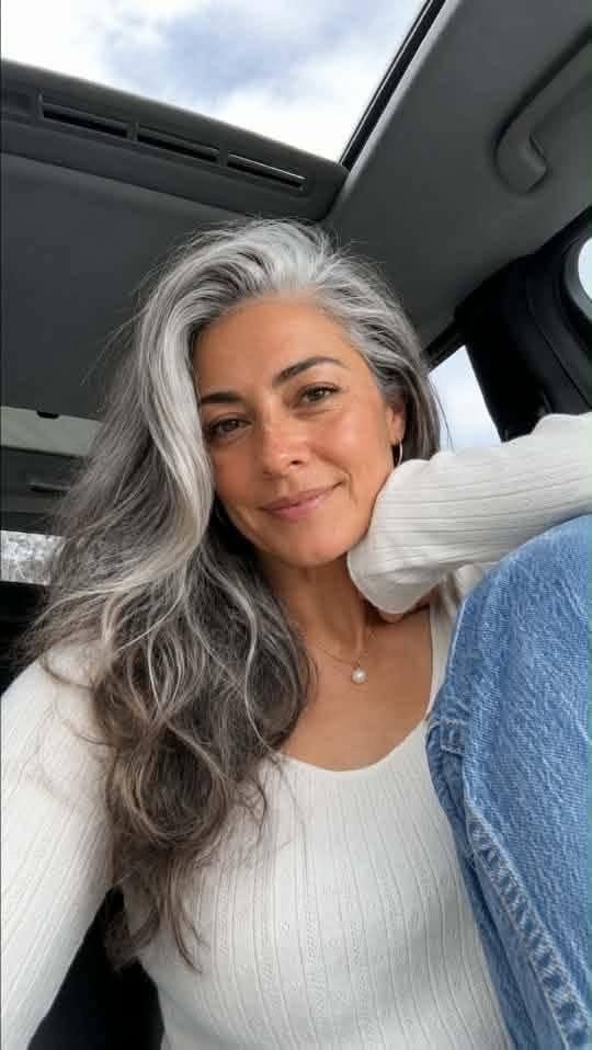 Woman with long silver-gray hair and dark roots smiling in a car