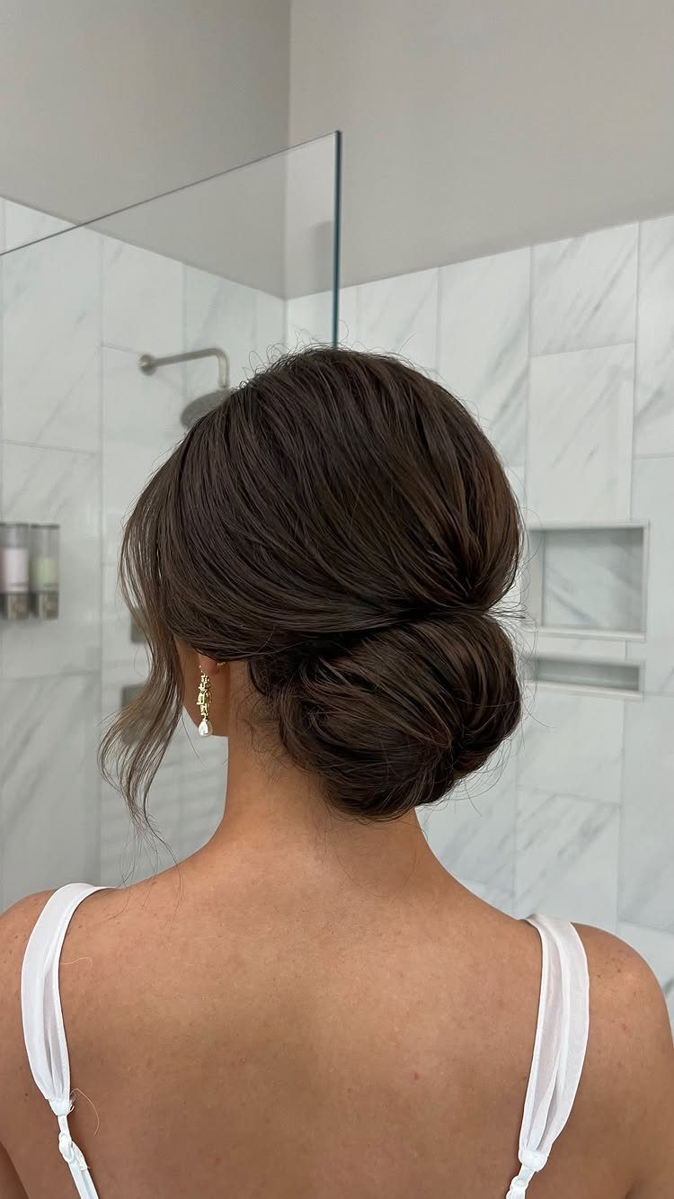Low chignon bun with loose face-framing strands against marble bathroom walls
