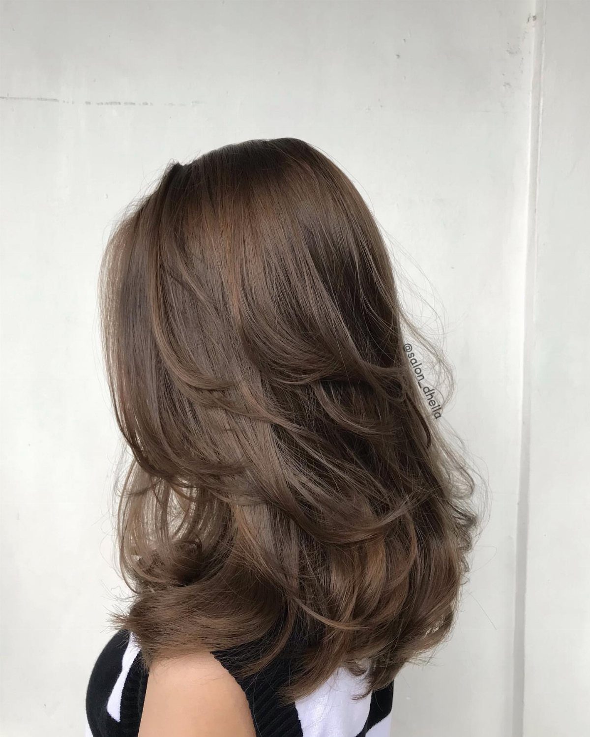 Shoulder-length brown hair falls in glossy, loose layered waves against a white wall