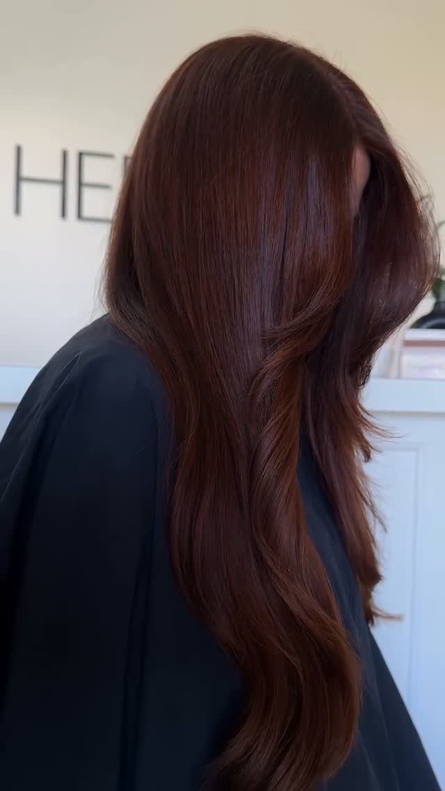Long, glossy deep auburn hair falls in loose waves over a dark salon cape