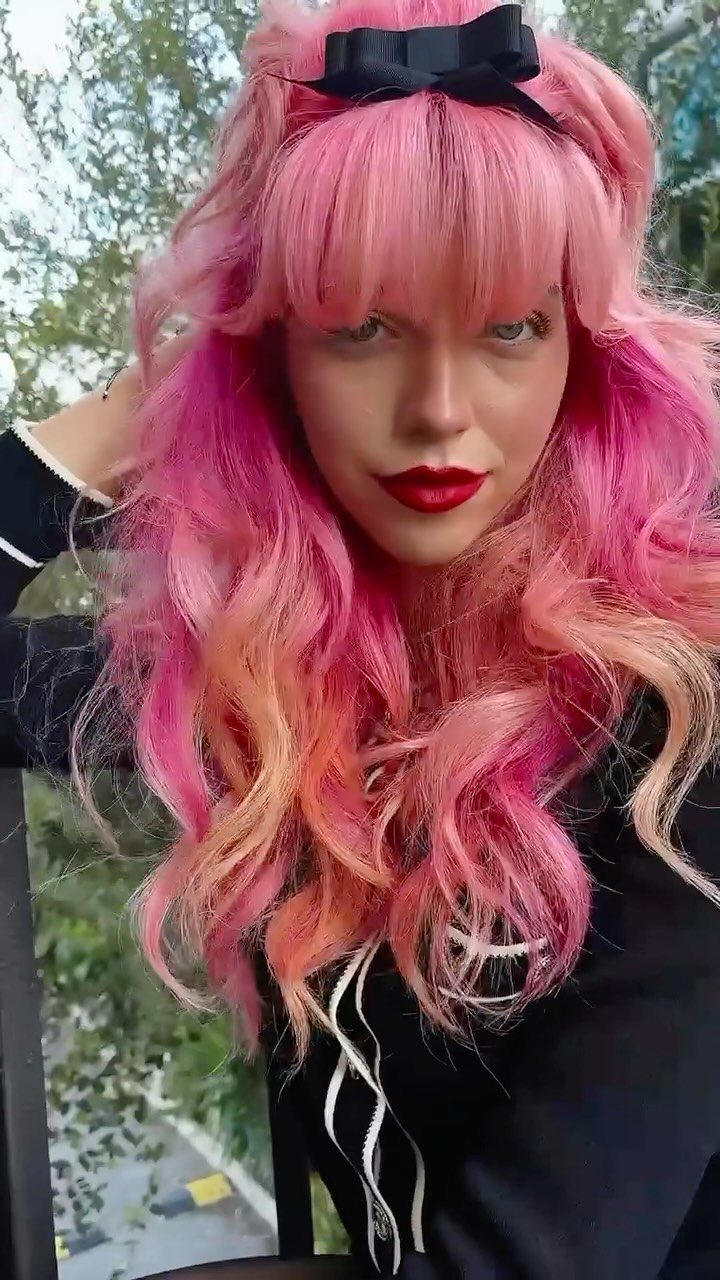 Long pink hair blends into peachy blonde ends beneath a black bow