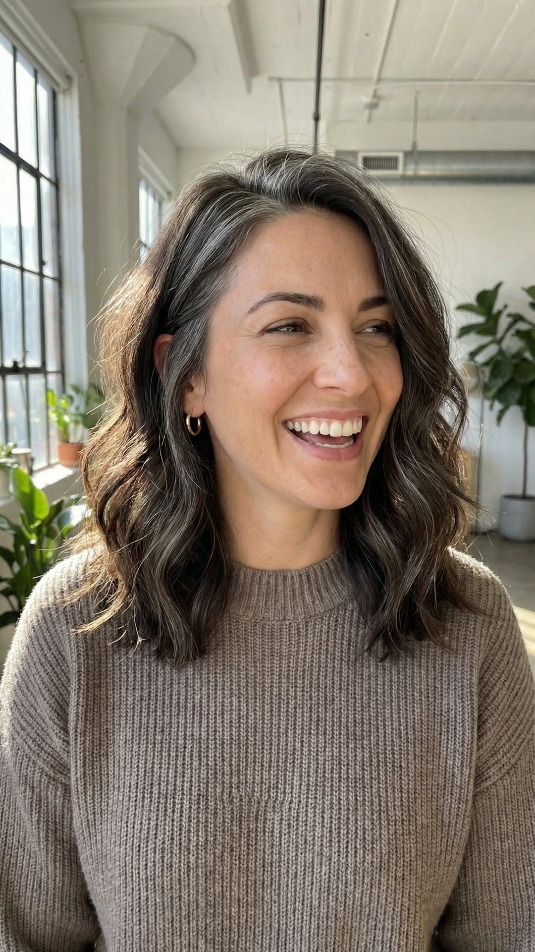 Woman with dark wavy hair showing silver-gray roots in a bright room