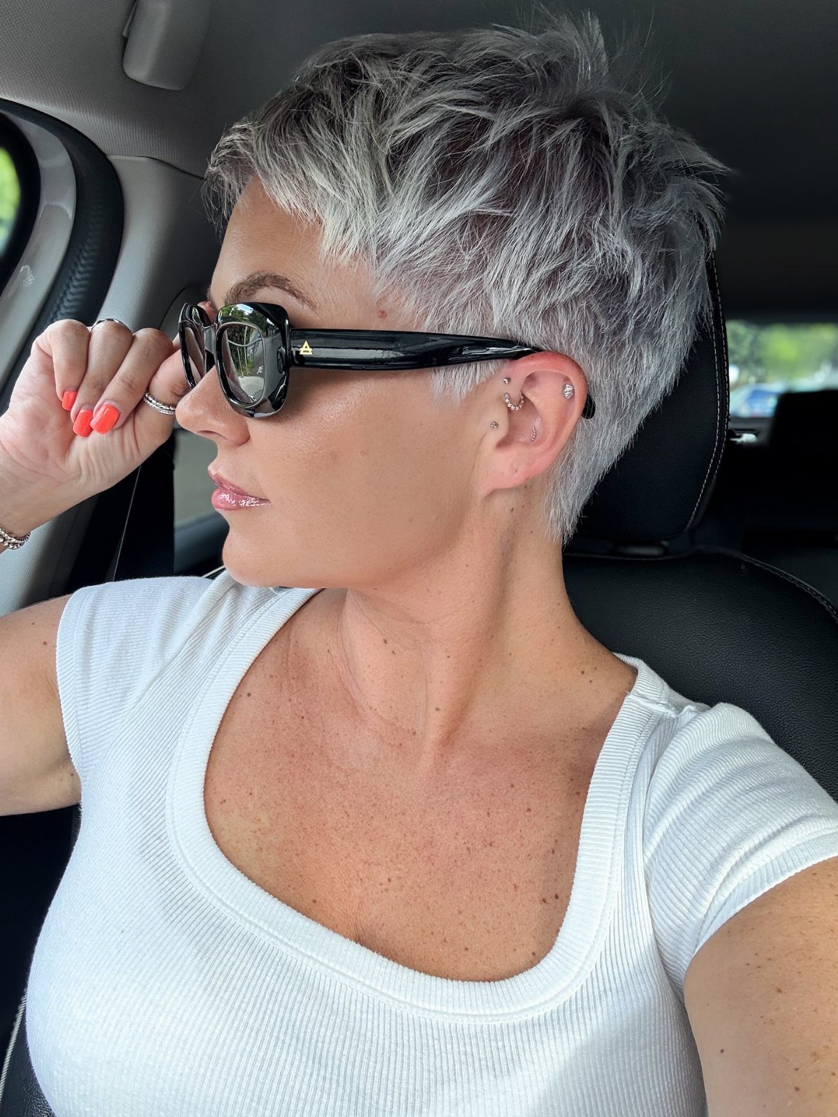 Woman with a short, tousled silver-gray pixie cut adjusting her sunglasses in a car