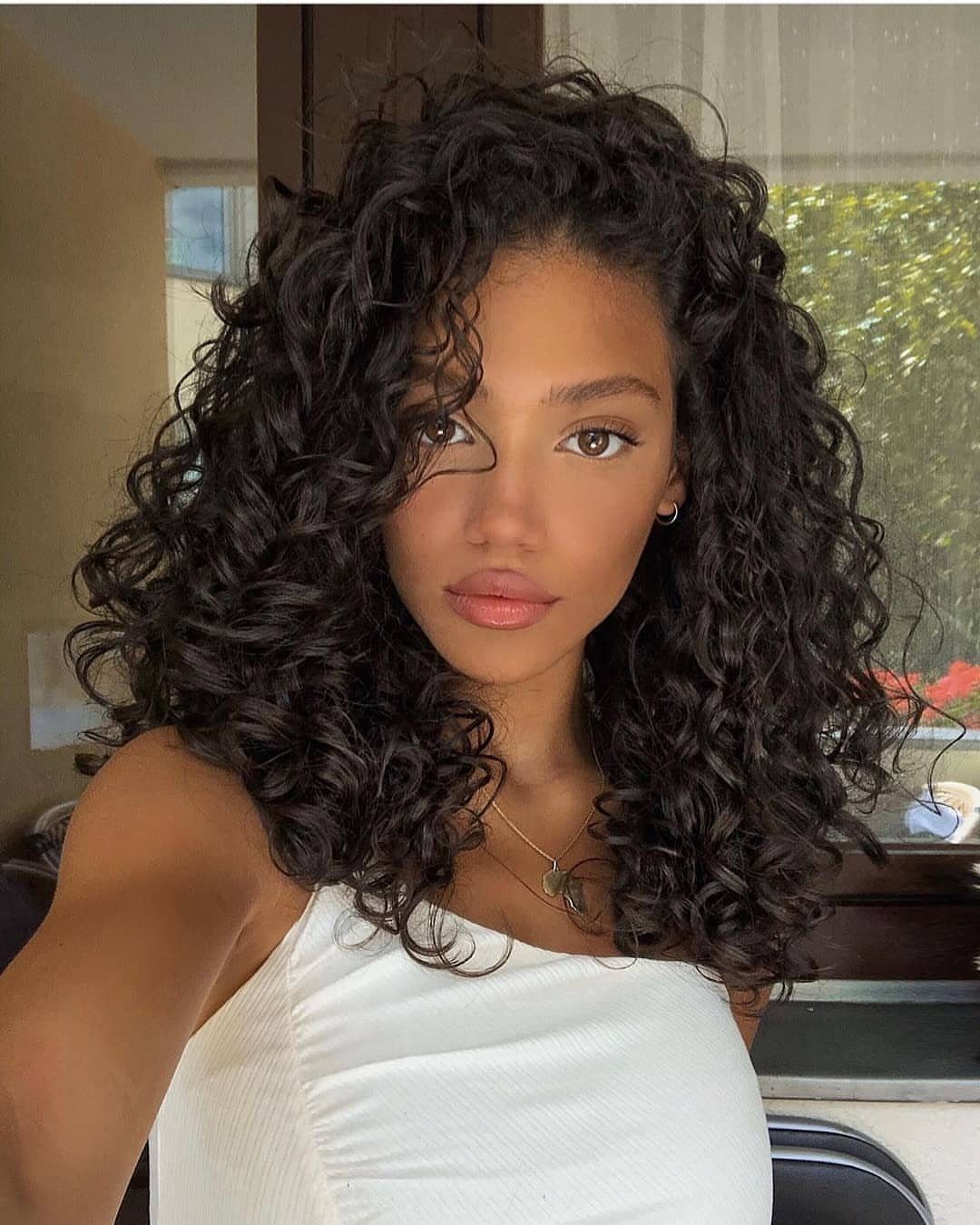 Woman with glossy black curls posing indoors in a white sleeveless top
