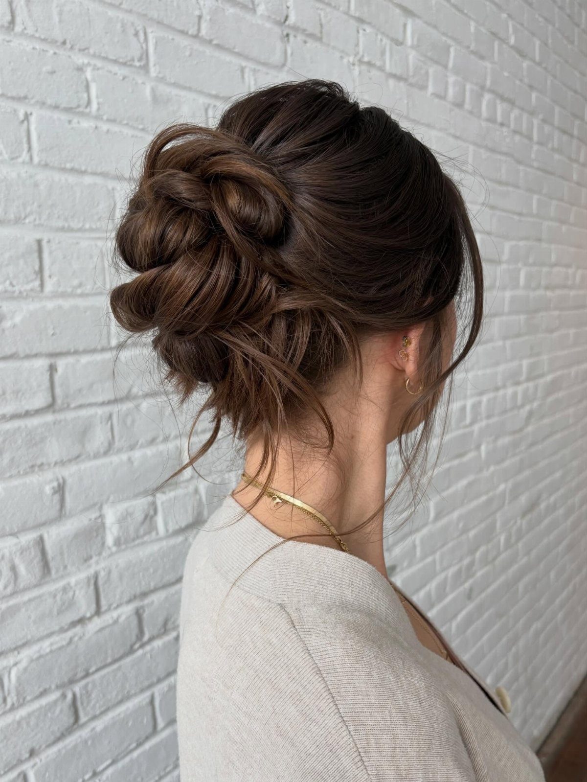 Side bun with twisted brown hair and loose wisps against a white brick wall