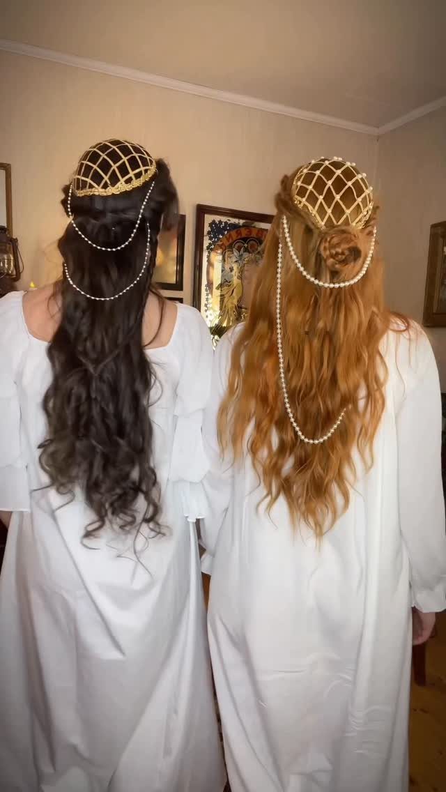 Two women with long hair wear pearl-draped gold hairpieces over white robes
